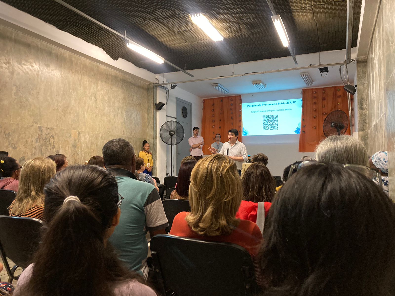 Na foto, o professor Marcel Haiaratsuka introduz o assunto da palestra e explica de onde surgiu a ideia do projeto. No palco estão outros dois professores, e no espaço estão os atendidos.